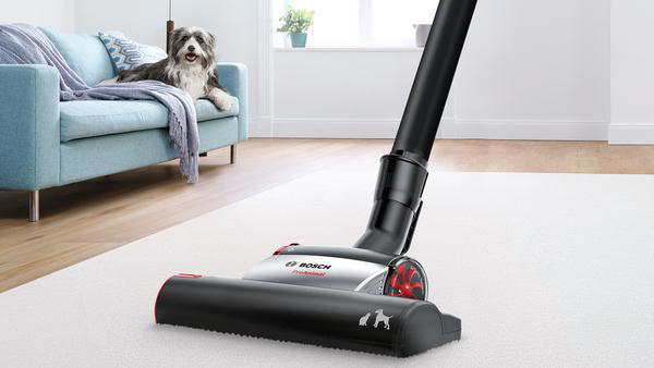 Ein Staubsauger auf einem weißen Teppich mit einem Hund auf einem Sofa im Hintergrund.