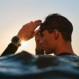 Mann im Wasser, der seine Schwimmbrille anpasst, mit Smartwatch und Sonnenaufgang im Hintergrund.