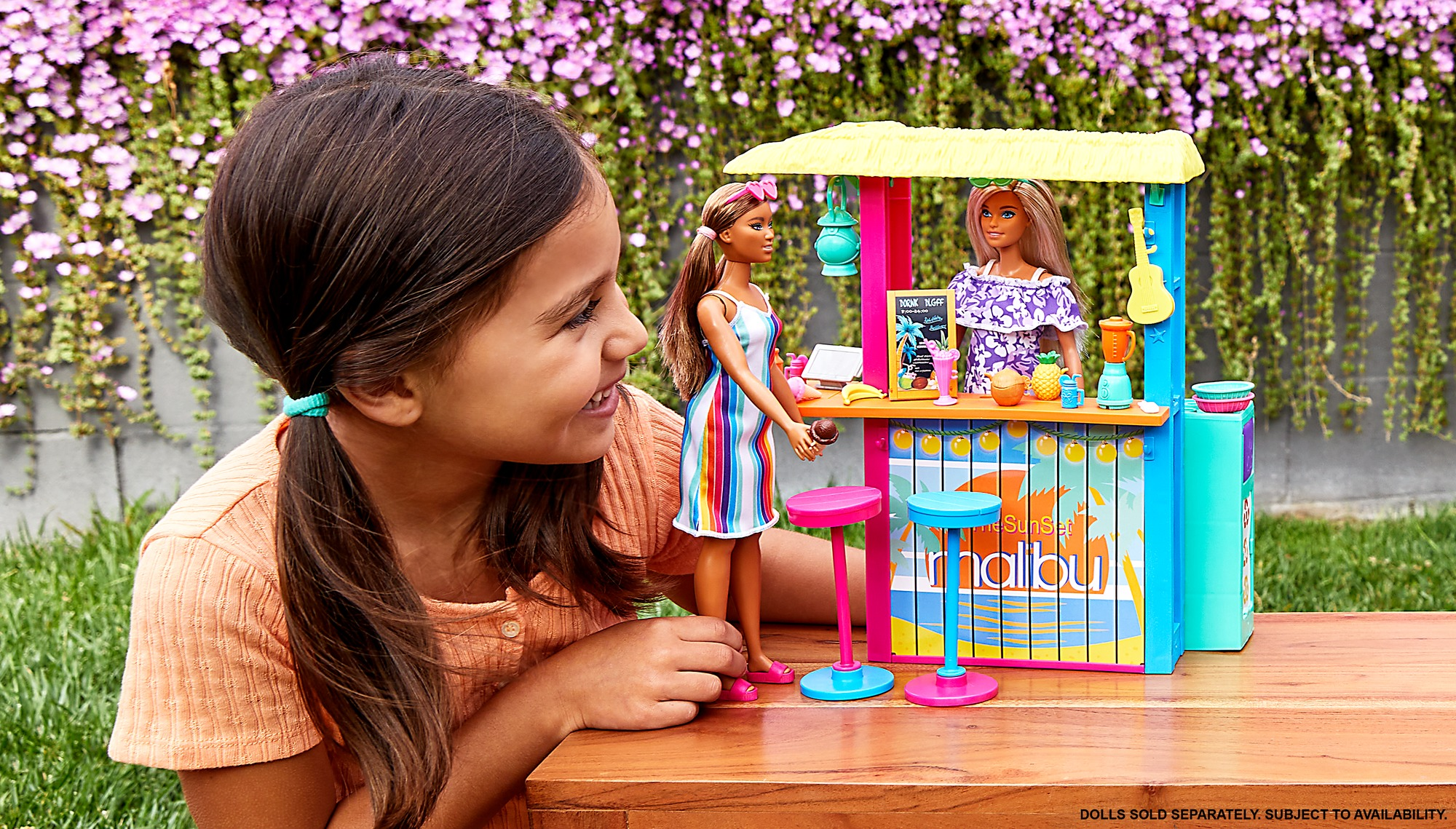 Ein Mädchen lächelt ein Barbie-Spielset an. Zwei Barbie-Puppen stehen hinter einer Malibu-Bar.