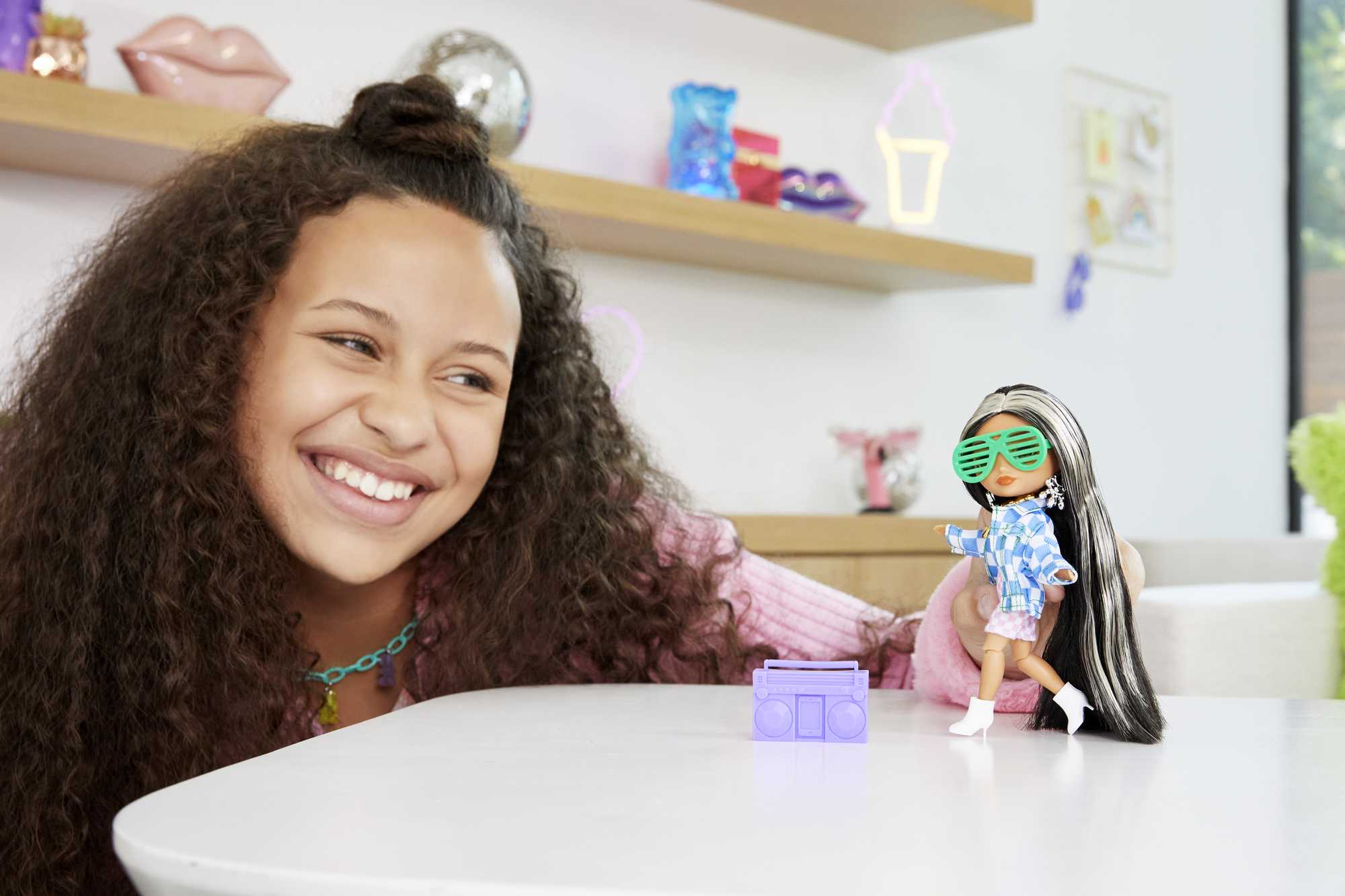 Ein junges Mädchen mit einer Puppe und einer Spielzeug-Boombox auf einem Tisch, lächelnd.