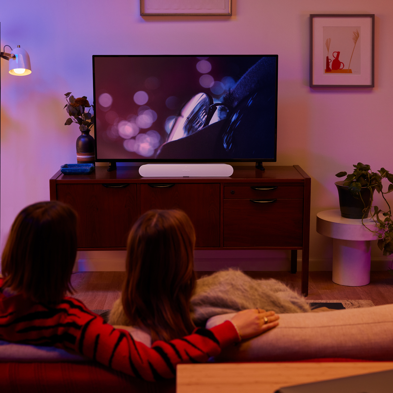 Woonkamerscène. Tv op een kast, soundbar eronder. Twee mensen op een bank kijken tv.