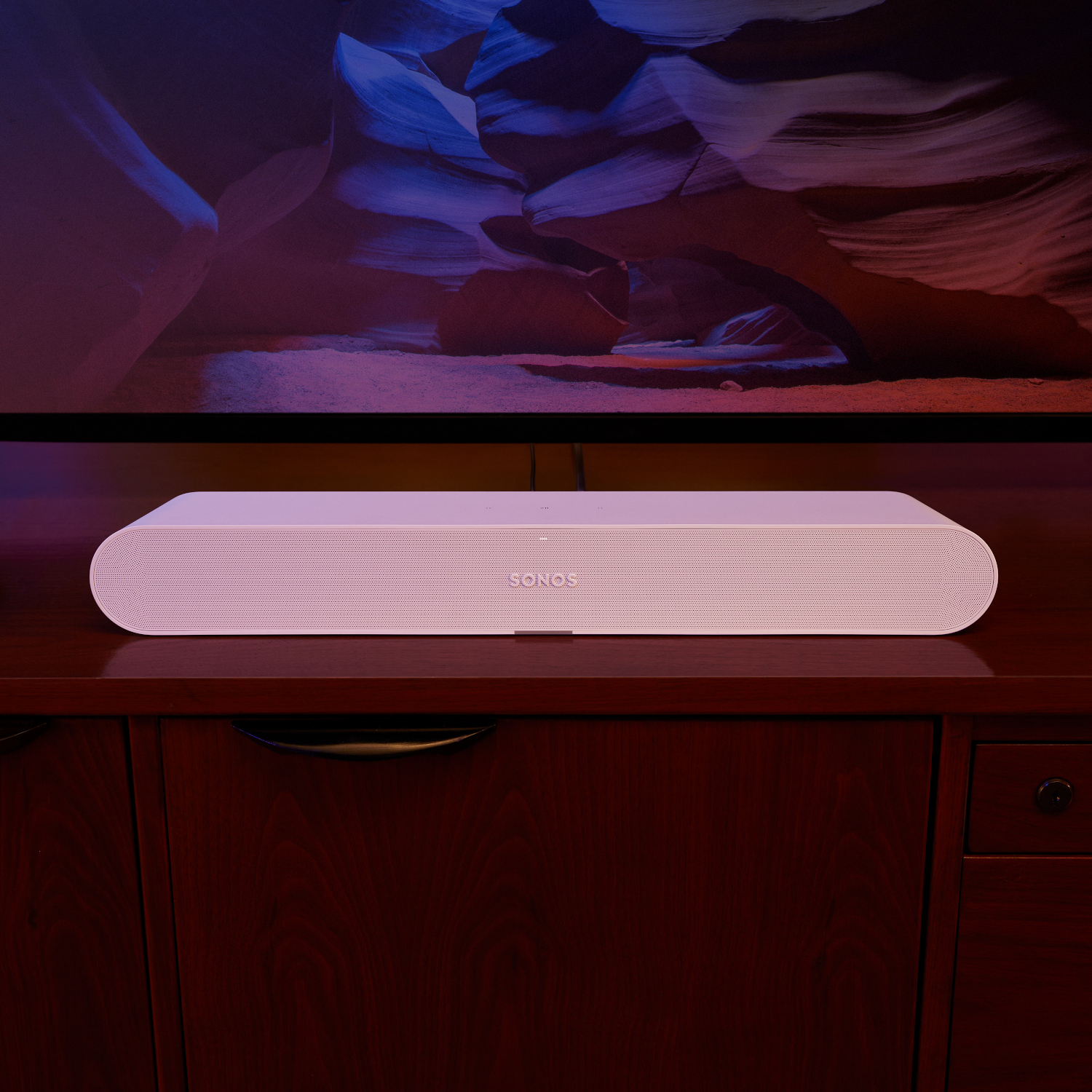 Een witte soundbar op een houten kast voor een verlicht tv-scherm.