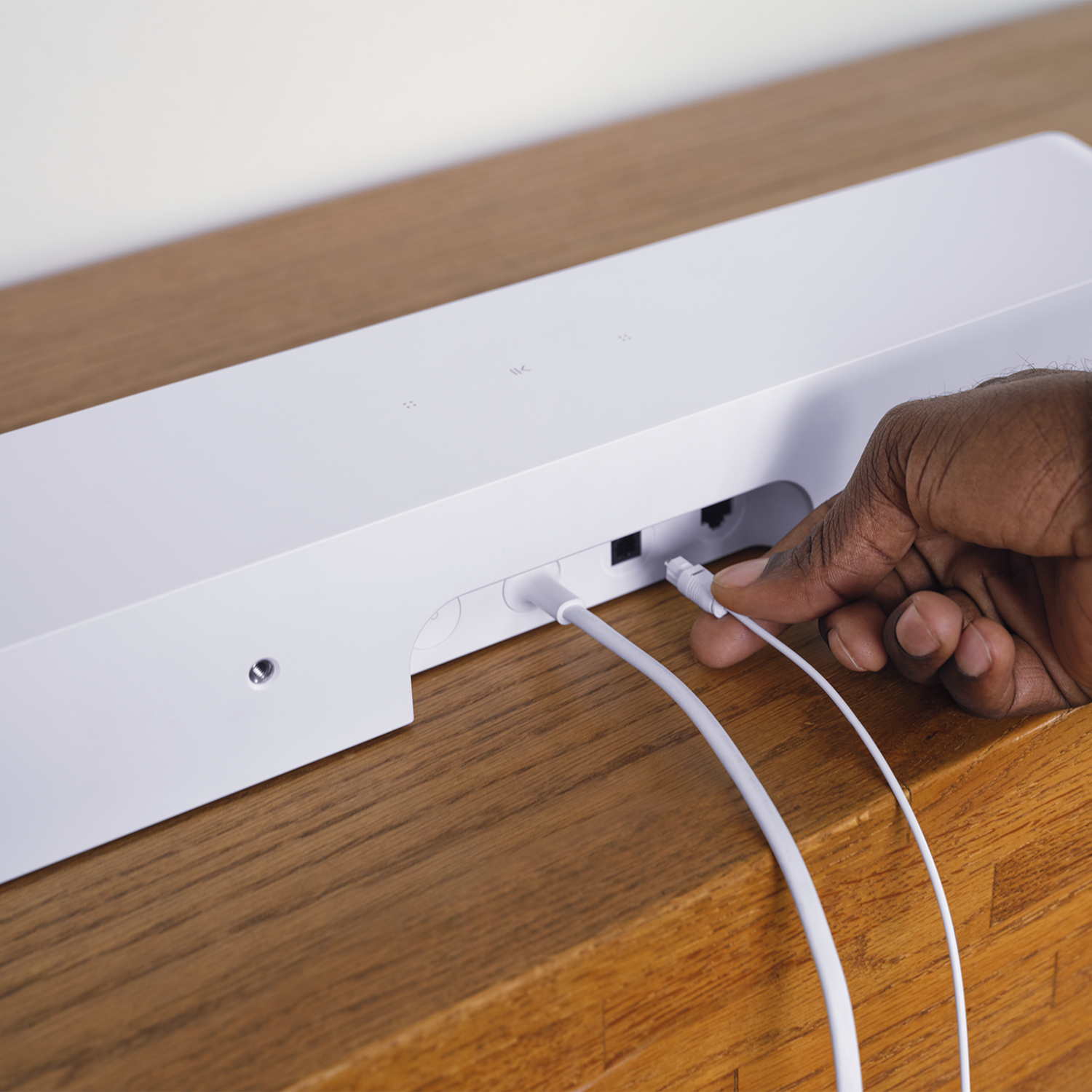 Een hand steekt een witte kabel in een witte soundbar. De soundbar staat op een houten oppervlak.