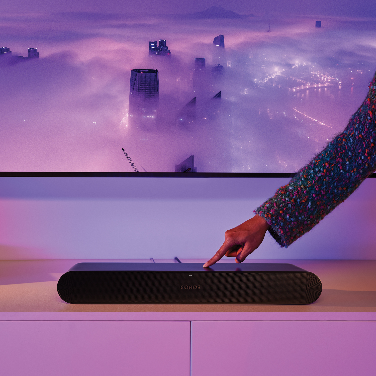 Hand raakt een zwarte Sonos soundbar aan op een wit oppervlak. Vage achtergrond met stadsbeeld.