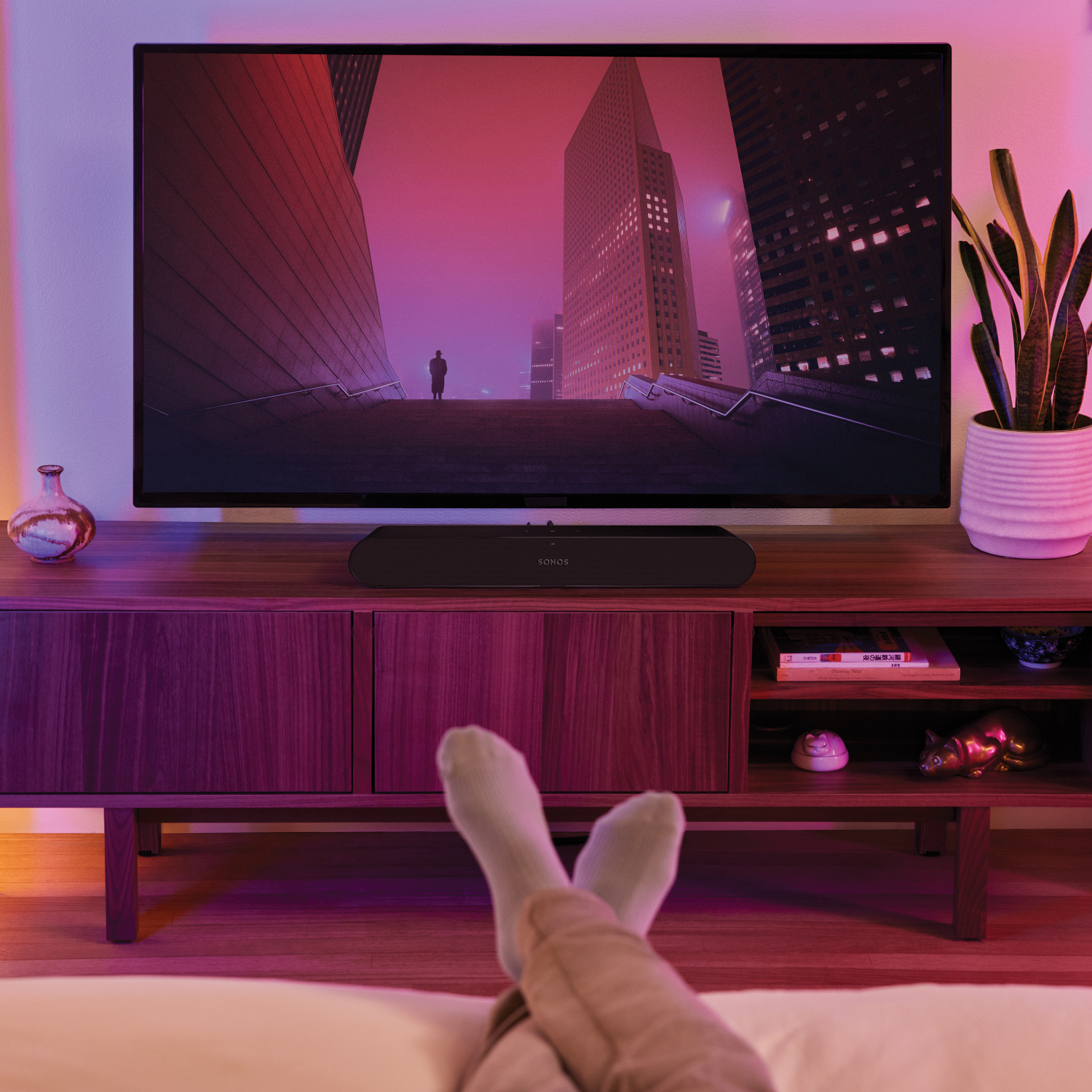 TV op een houten kast, benen gekruist. TV toont een stadsgezicht. Soundbar eronder.