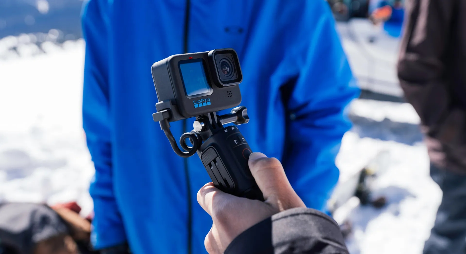 Eine Person hält eine GoPro-Kamera auf einem Stock vor verschneitem Hintergrund.