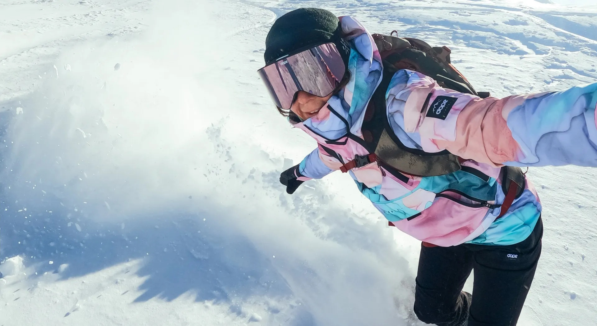 Person beim Snowboarden, mit Schnee bedeckt. Pinke und blaue Jacke mit Brille. Schnee fliegt um die Person herum.