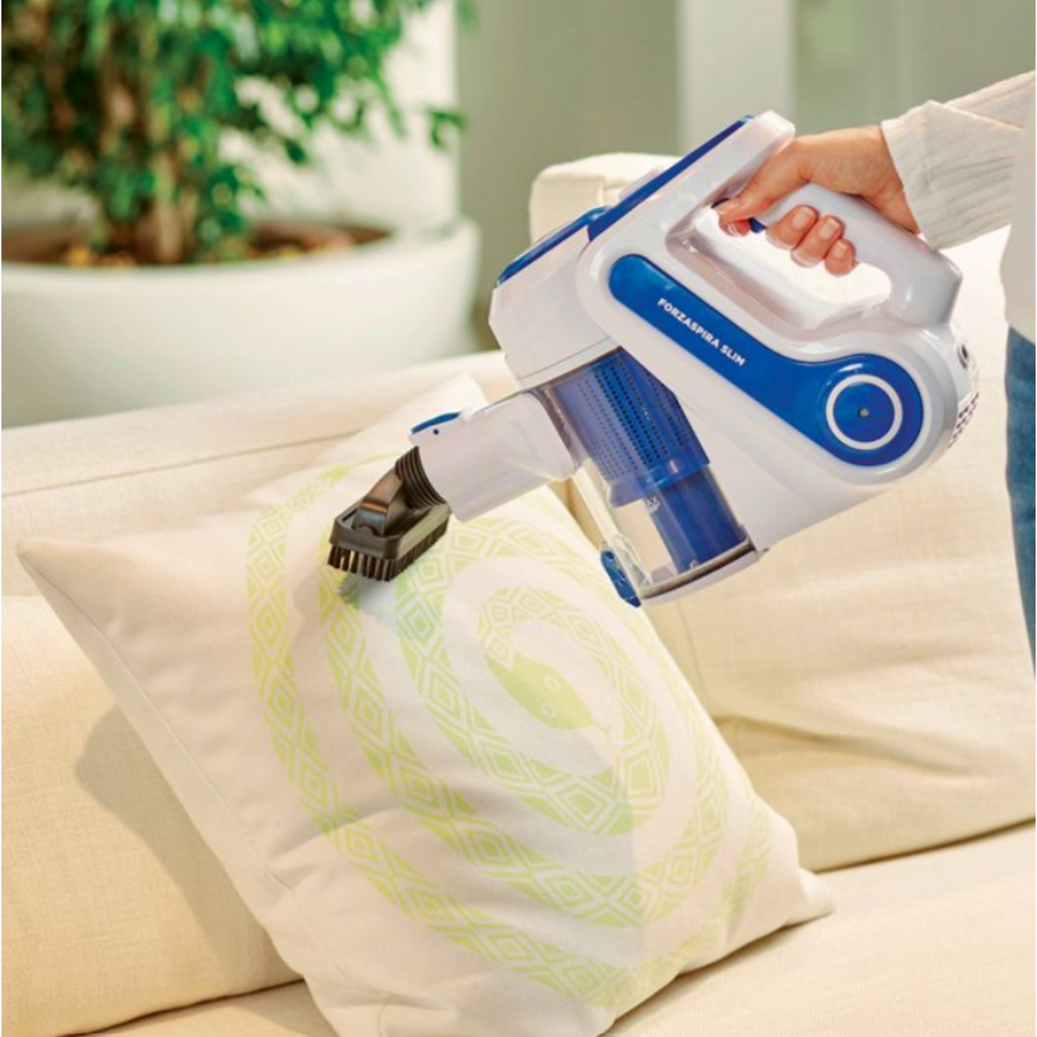 Una aspiradora de mano limpia una almohada. La aspiradora es blanca y azul. La almohada es blanca con un patrón verde.