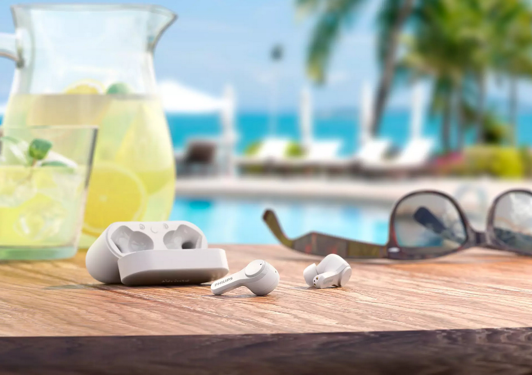 Weiße Ohrhörer mit Ladecase auf Holzoberfläche. Limonade, Sonnenbrille und Meer im Hintergrund.