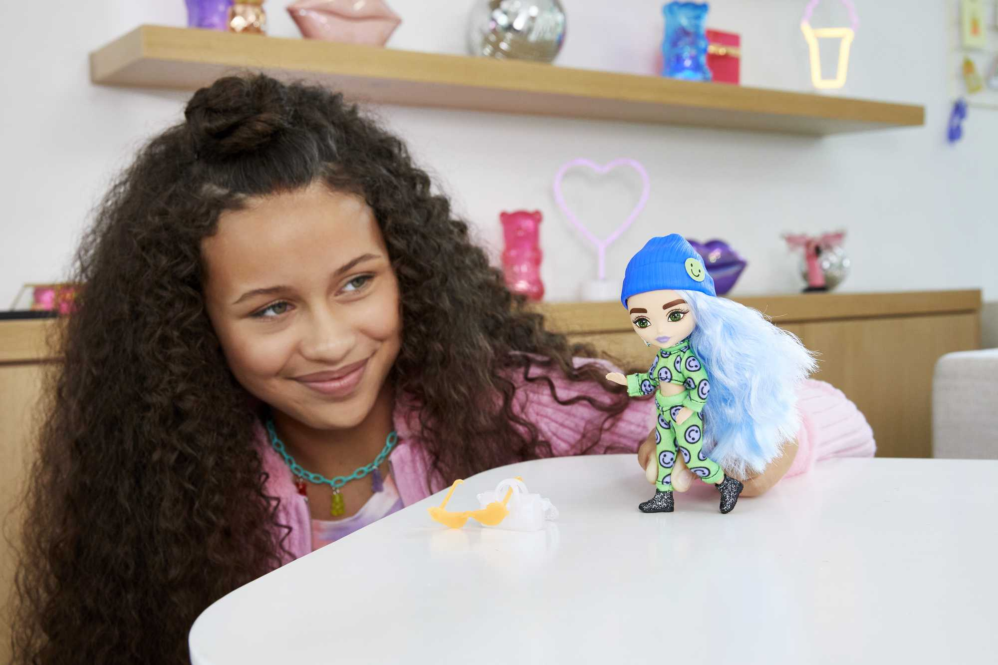 Ein junges Mädchen lächelt eine Puppe an, die auf einem Tisch sitzt. Die Puppe hat blaue Haare und einen grünen Anzug.