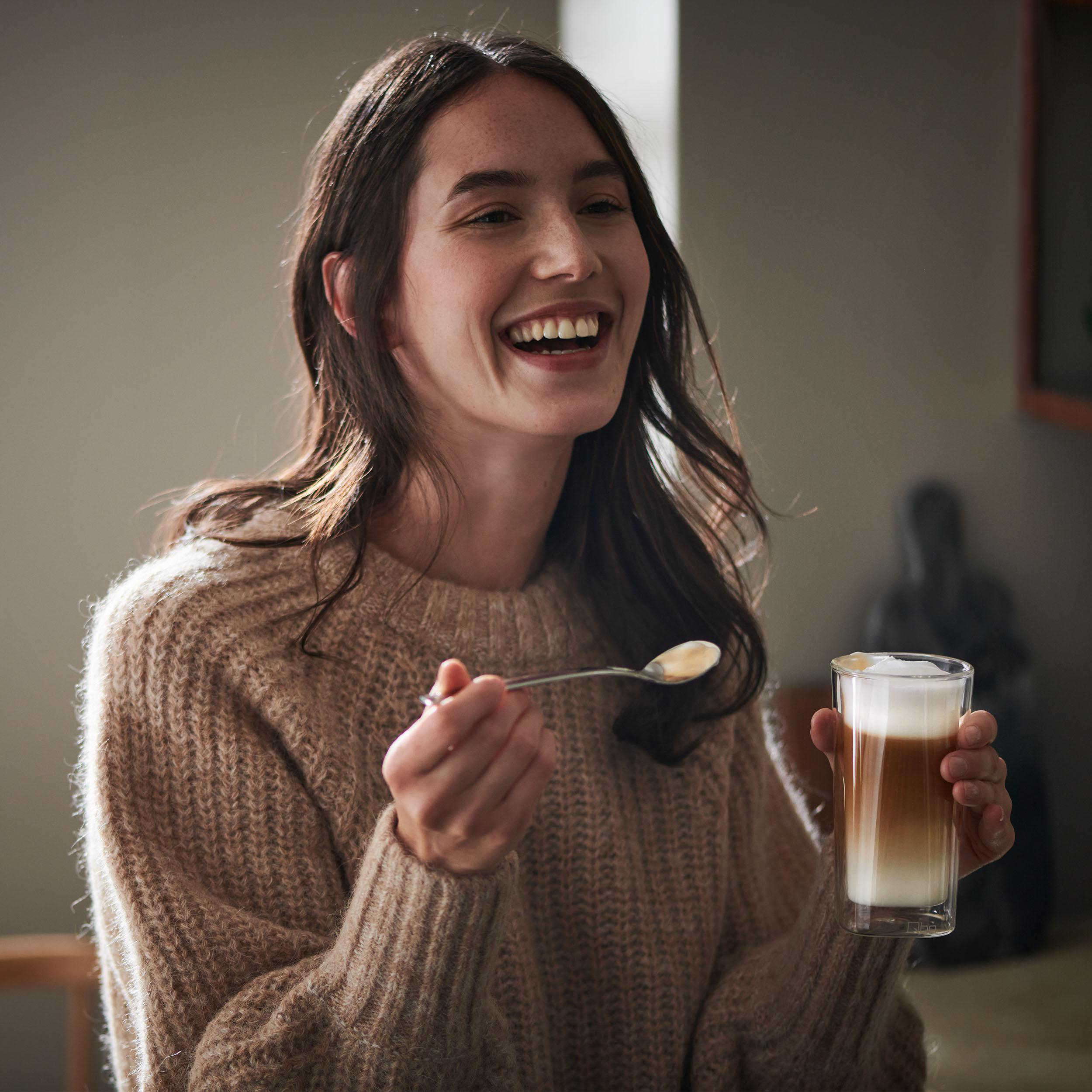 Lachende Frau, die einen Löffel und ein Glas Latte mit Schaumschicht hält.