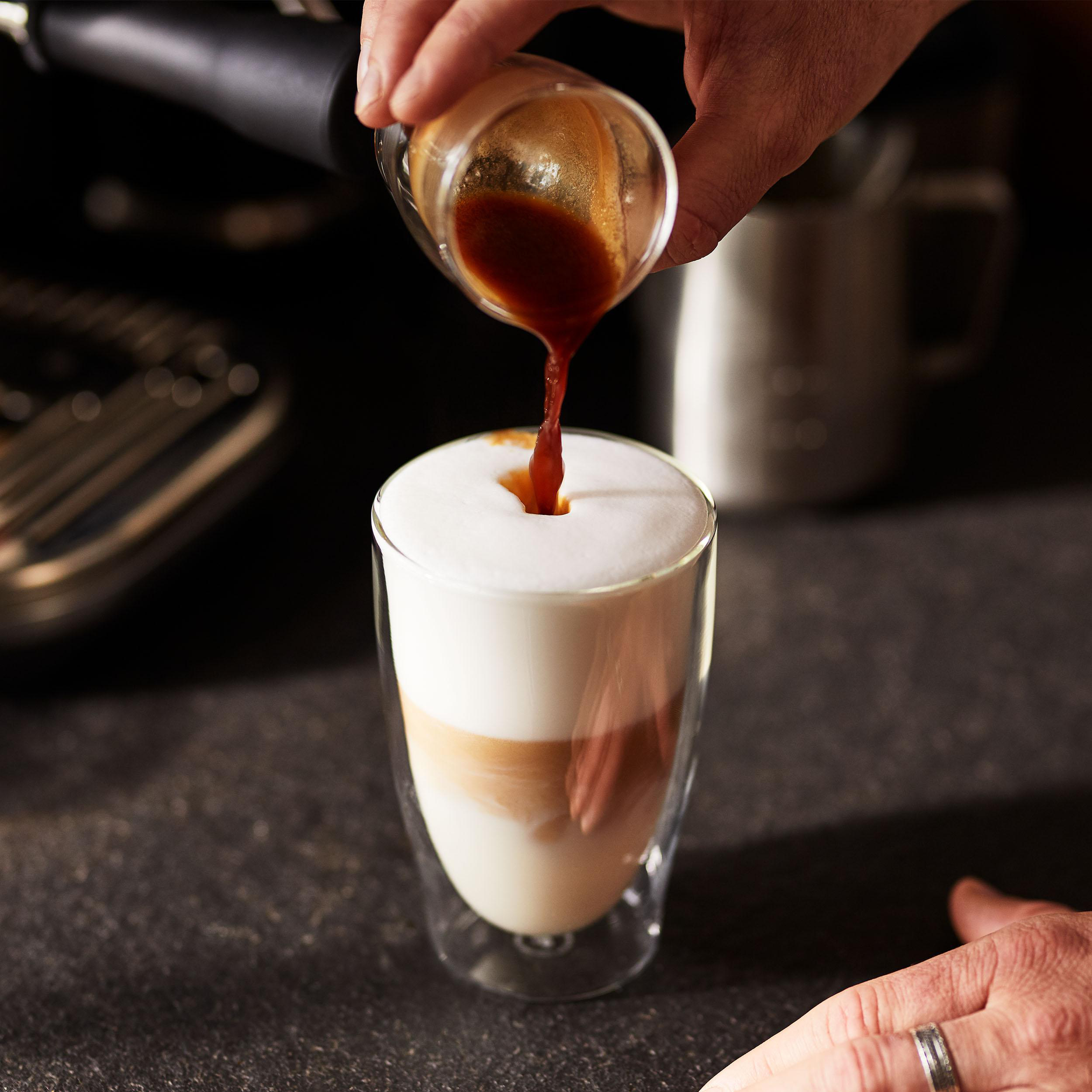 Eine Hand gießt Espresso in ein Glas geschichteten Latte, mit Schaum oben drauf. Andere Hand im Bild.