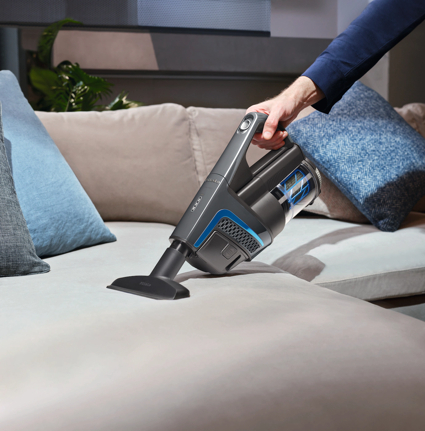 Une main utilise un aspirateur sur un canapé. L'aspirateur est gris et bleu. Le canapé est beige.