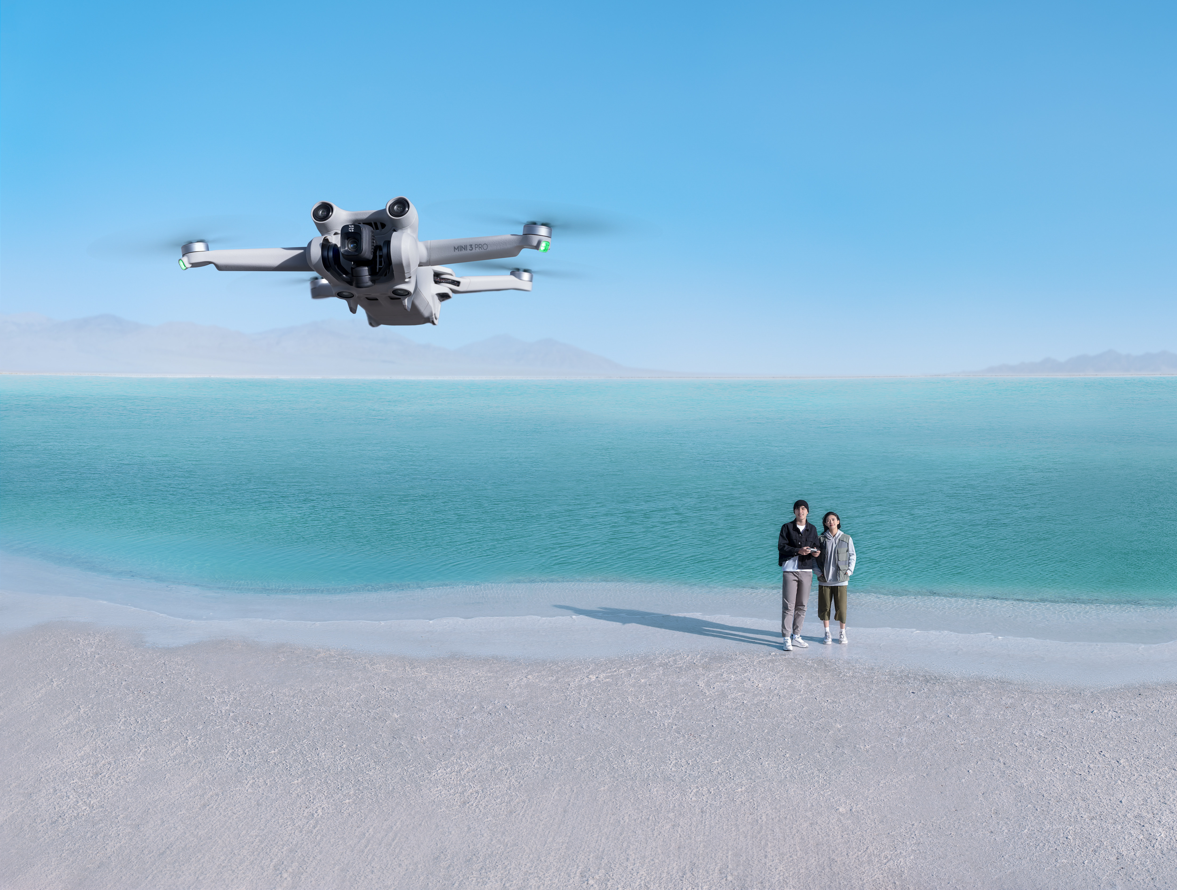 Drohne im Flug über einem Strand mit zwei Personen. Blauer Himmel und Ozean im Hintergrund.