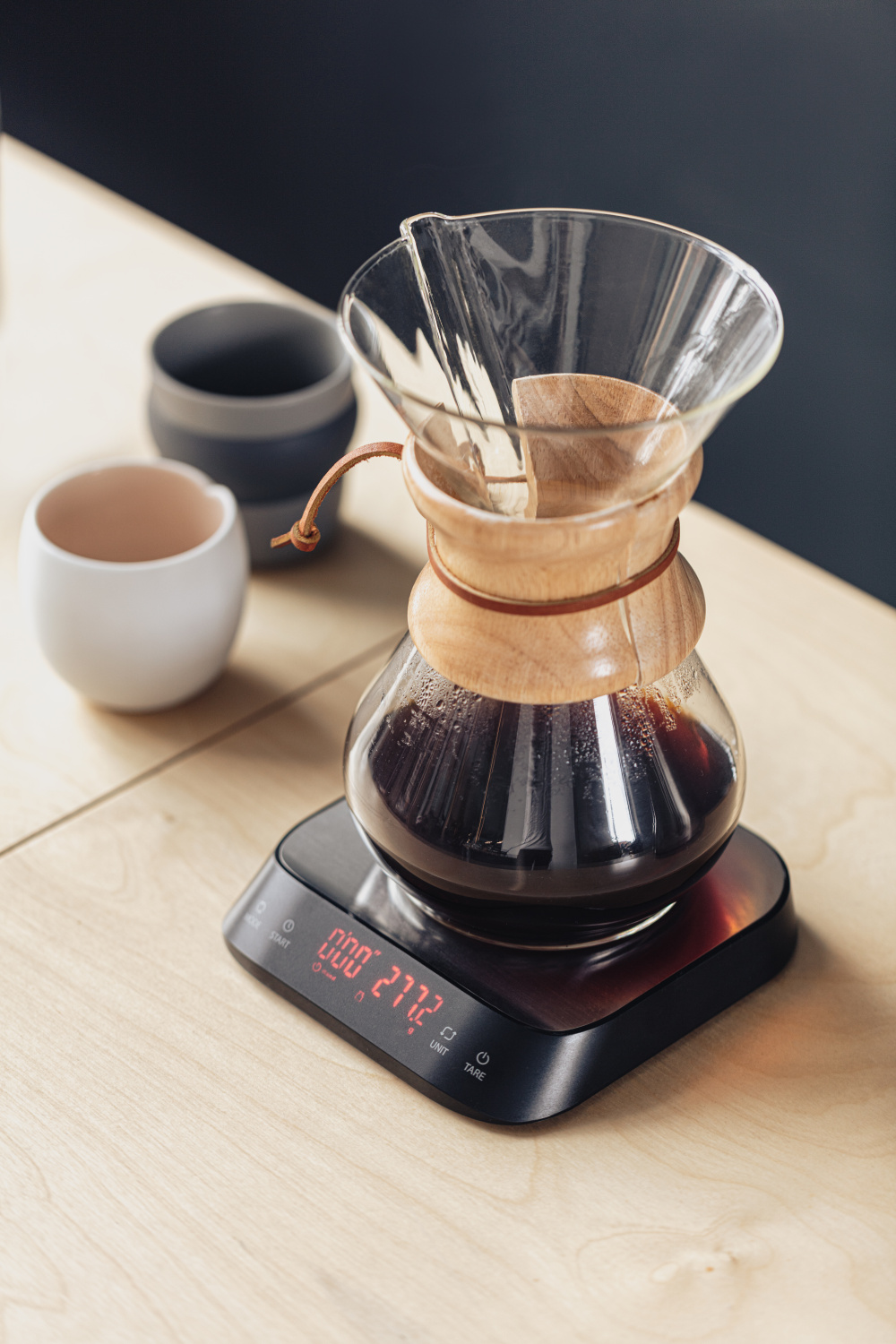 Üveg kávéfőző mérlegen. Pour-over beállítás faasztalon, sötét háttérrel.