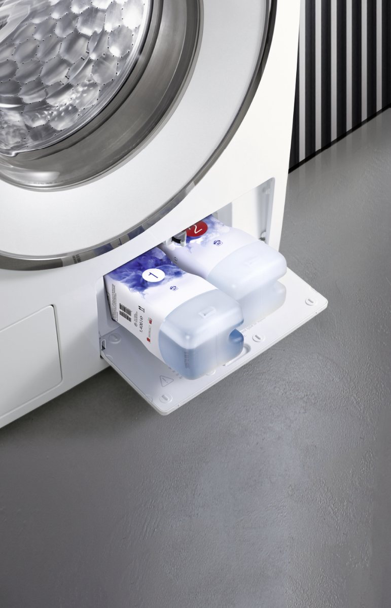 A washing machine drawer is open, with two detergent containers visible. The machine is white, with a gray floor.