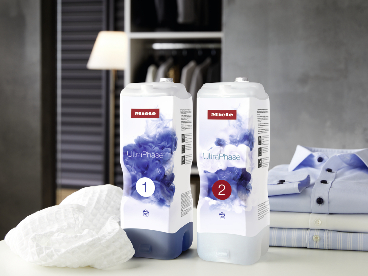Two Miele UltraPhase laundry detergent bottles, clothes on a table, and a wardrobe in the background.