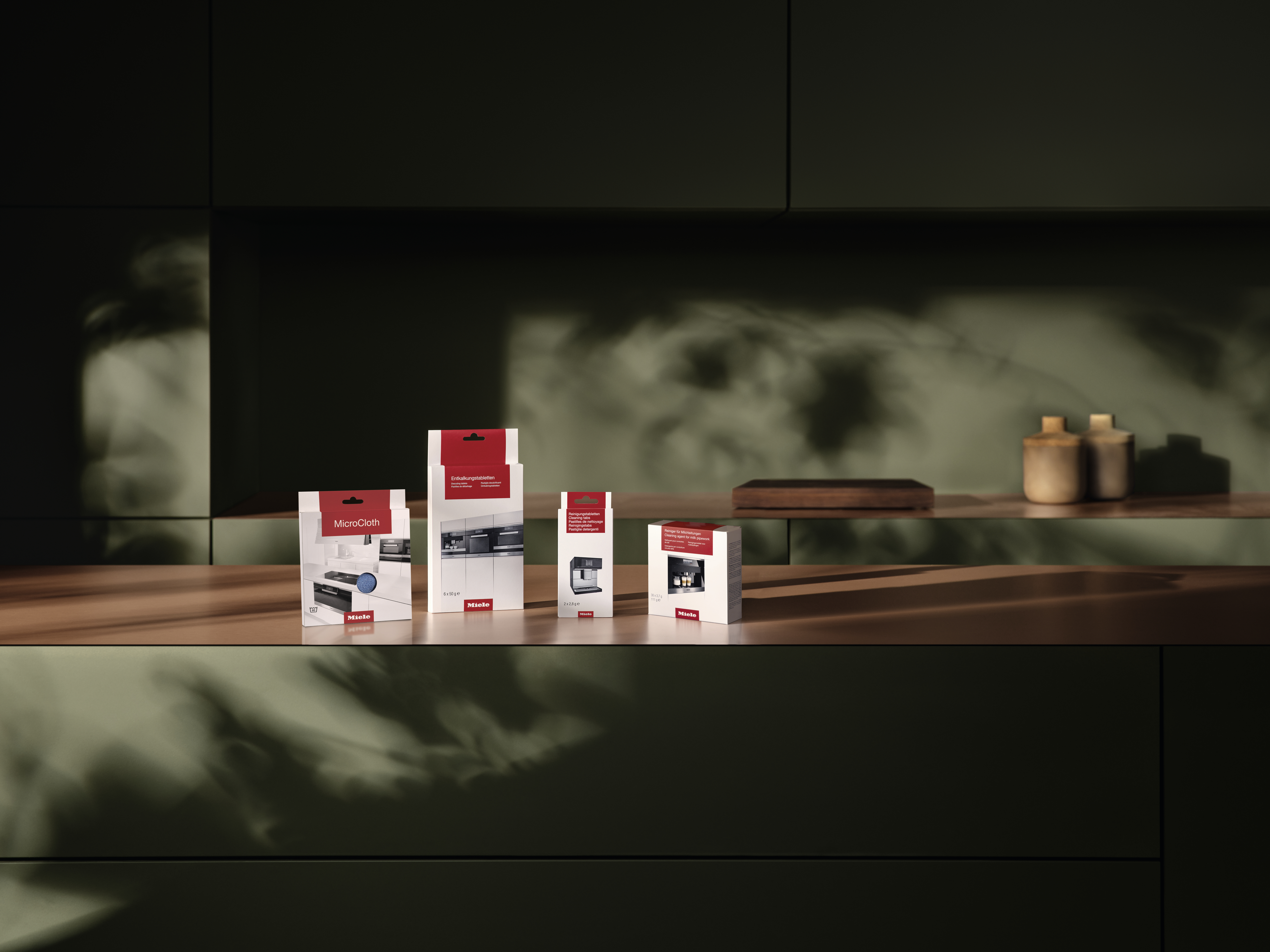 Schachteln auf einem Regal mit Miele-Produkten. Der Hintergrund ist eine dunkelgrüne Küchenumgebung.