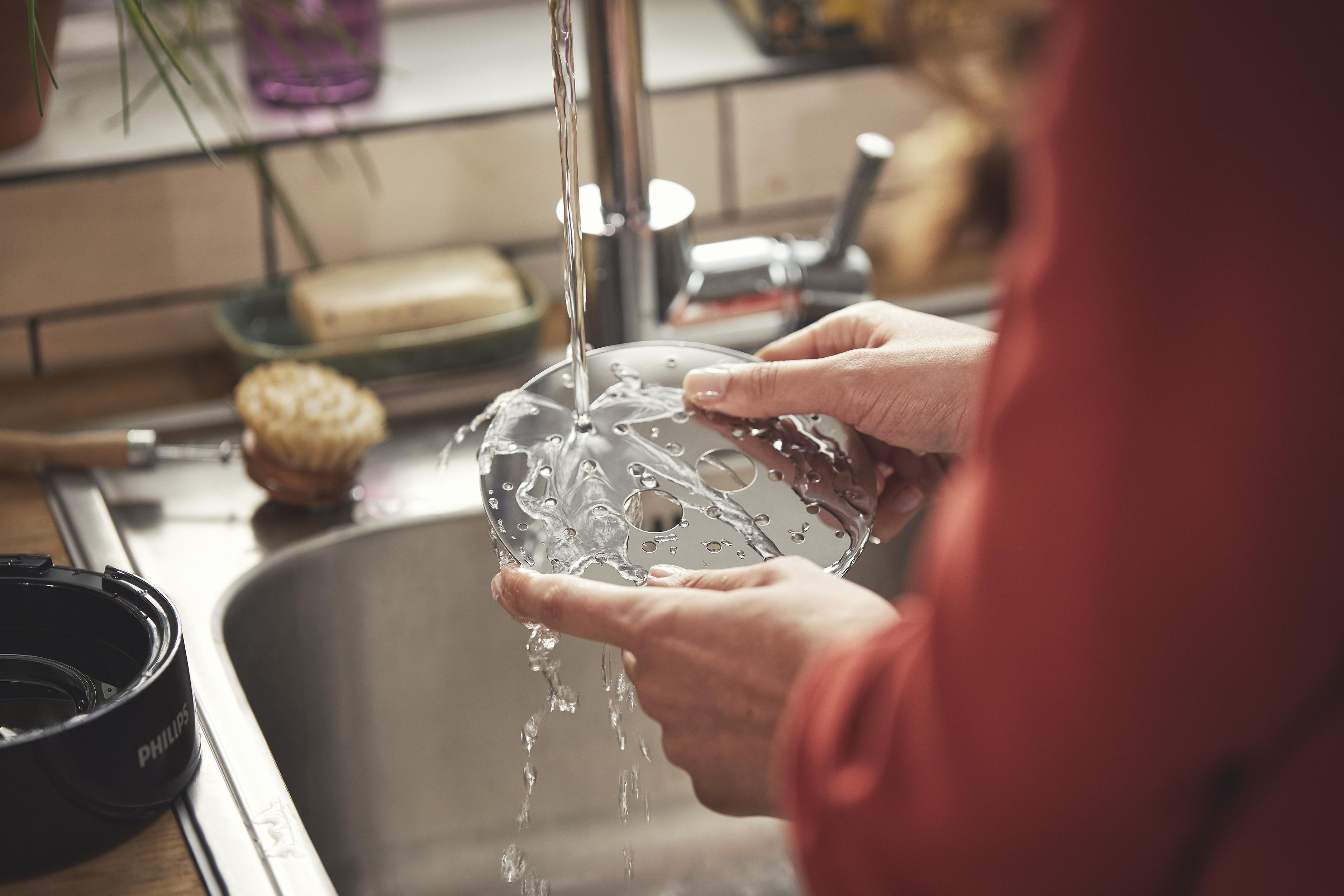 Hände waschen eine Küchenmaschine unter fließendem Wasser in einem Küchenbecken, andere Gegenstände sind sichtbar.