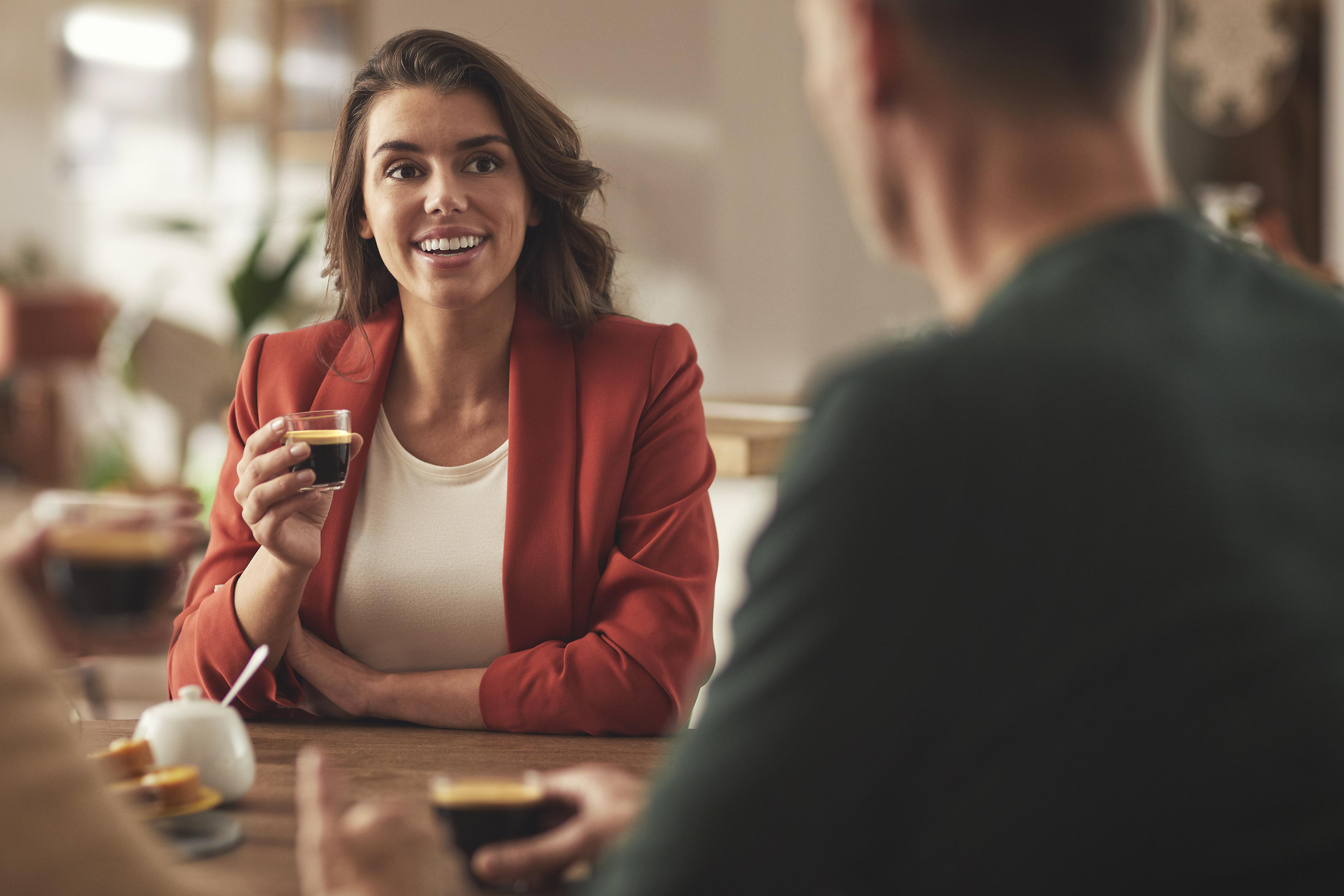Eine lächelnde Frau in einem roten Blazer hält ein kleines Glas mit dunkler Flüssigkeit.