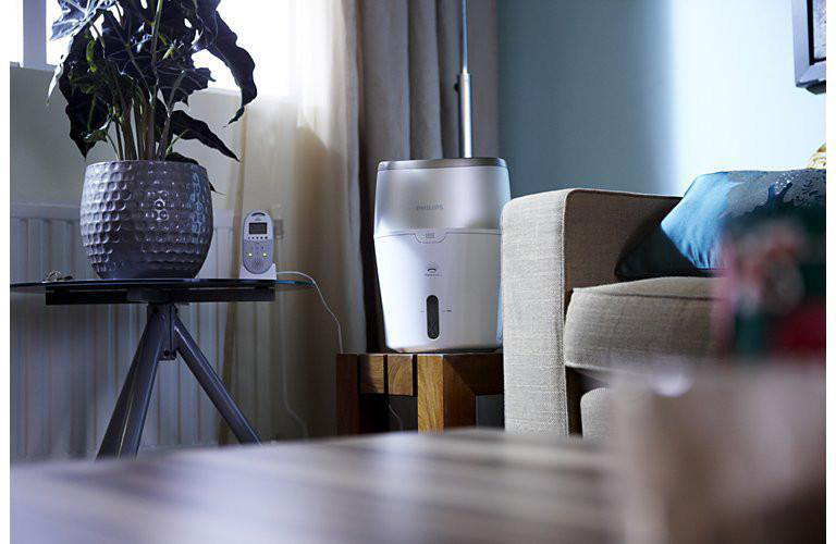 Wohnzimmer mit Luftbefeuchter auf einem Tisch. Eine Pflanze, ein Monitor und ein Sofa sind ebenfalls zu sehen.