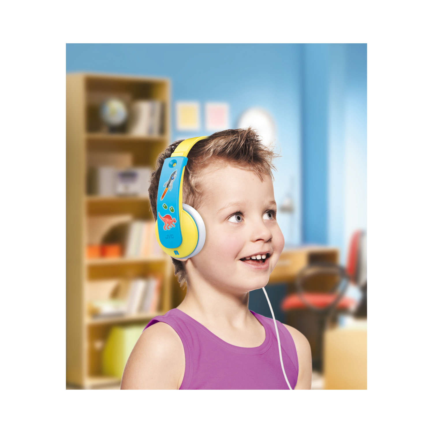 Un niño pequeño con auriculares azules y amarillos sonríe mientras mira hacia arriba, con una estantería de fondo.