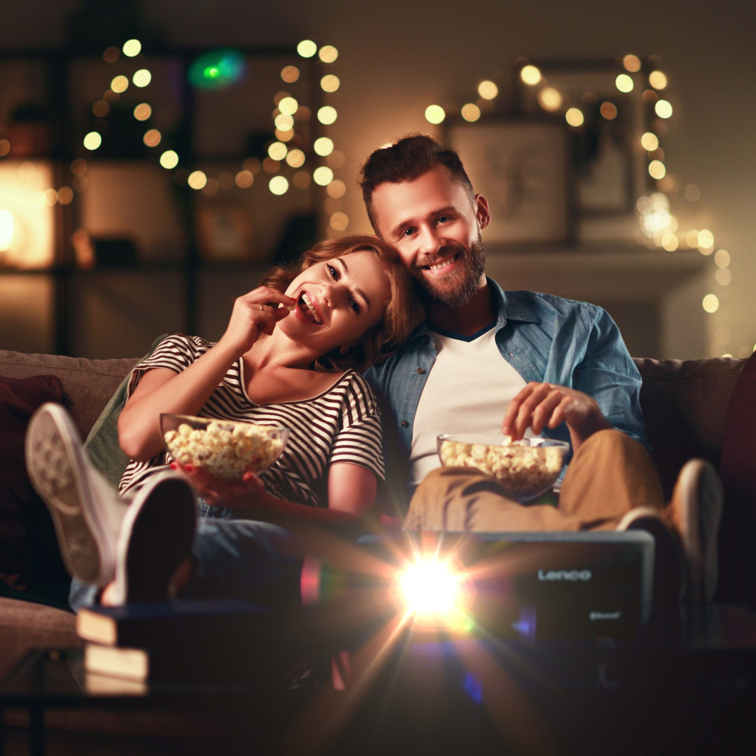 Couple sur canapé regardant film, mangeant pop-corn. Projecteur devant, lumières derrière.