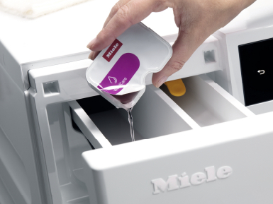 A hand pouring liquid from a white package into a white washing machine drawer.