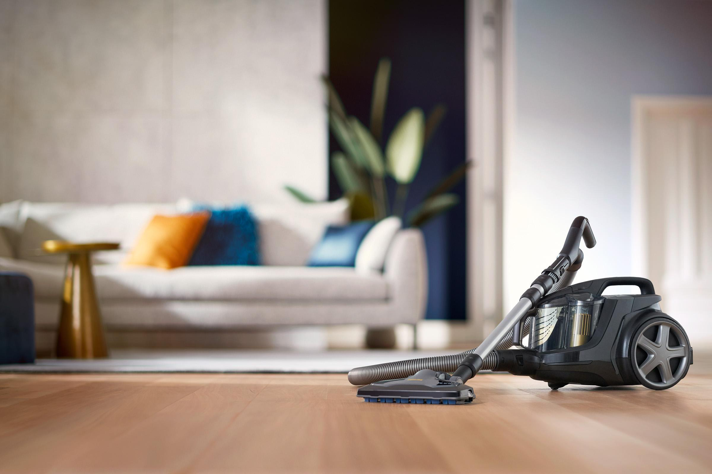 Ein Philips Staubsauger mit Düse auf Holzboden, mit Couch und Pflanze im unscharfen Hintergrund.