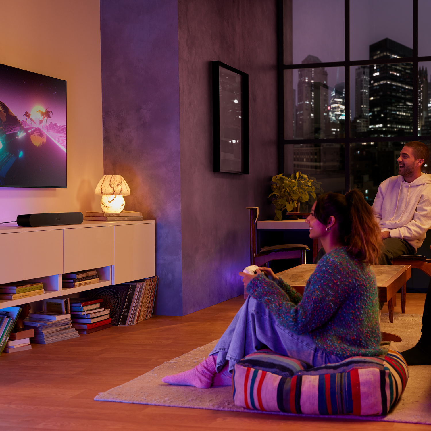 Dos personas en una habitación, viendo la televisión. Una lámpara, un armario y una ventana con vista a la ciudad son visibles.