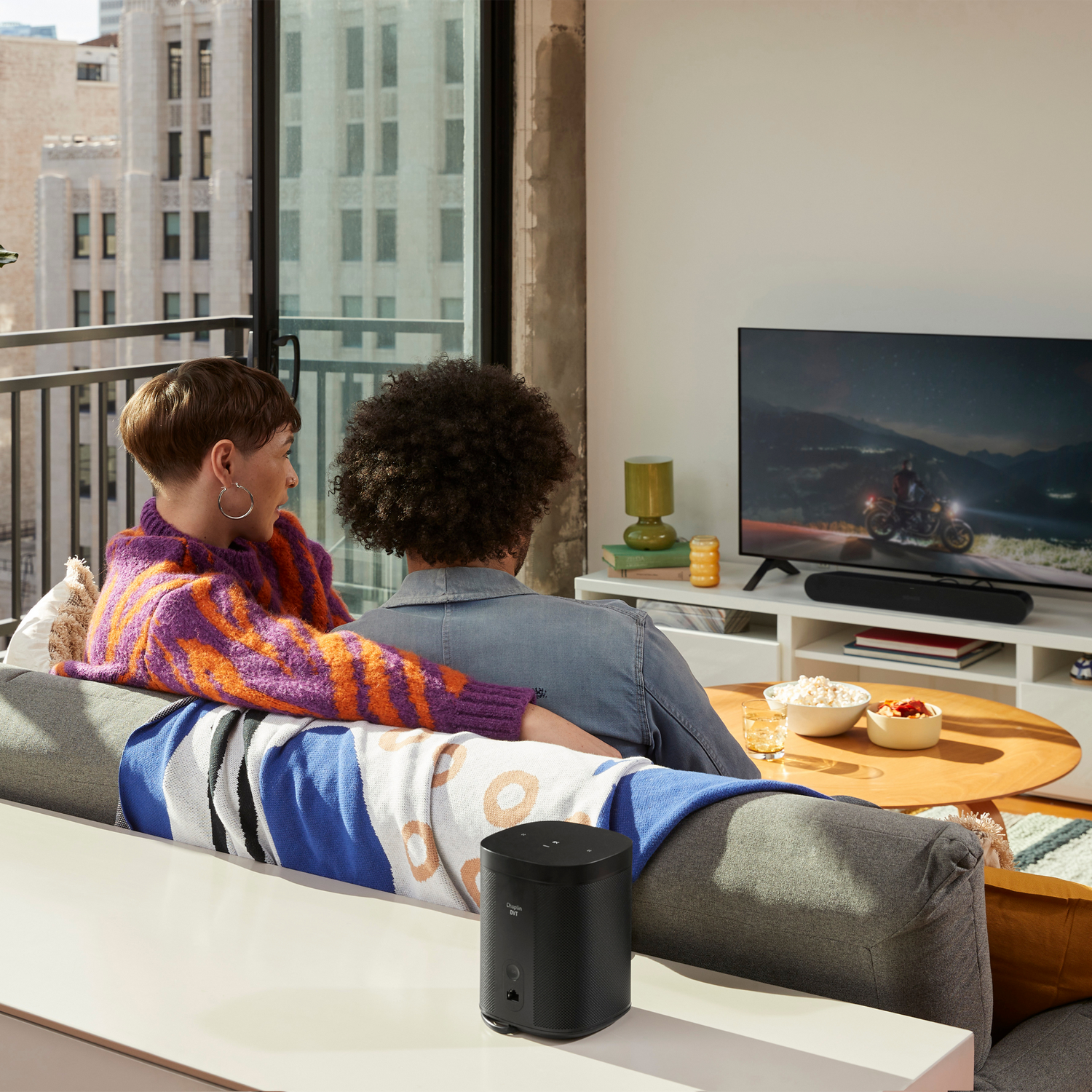 Dos personas en un sofá viendo la televisión, hay un altavoz frente a ellos y una ventana detrás.