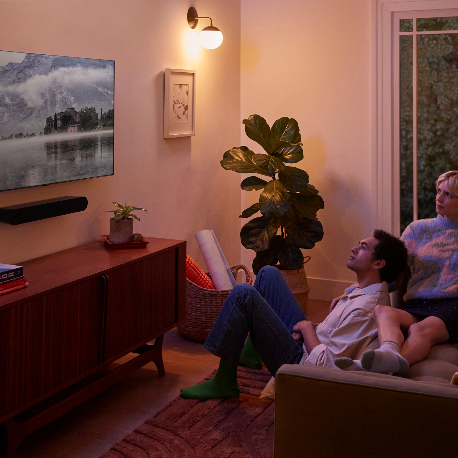 Dos personas en un sofá viendo la televisión. Un televisor está montado en la pared encima de un mueble y decoración.