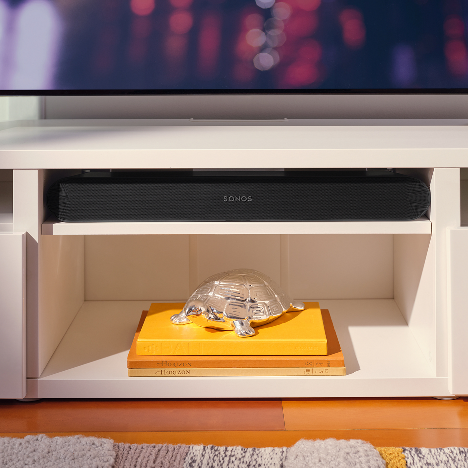 Una barra de sonido Sonos negra en una estantería blanca, con libros y un adorno de tortuga plateada. Ambiente de habitación.