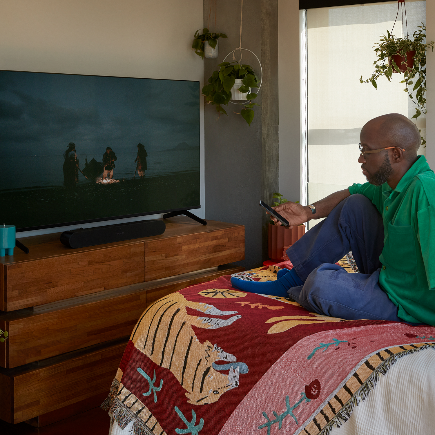 Un hombre está sentado en una cama, viendo la televisión. Una barra de sonido está en el gabinete debajo. Una manta estampada está en la cama.