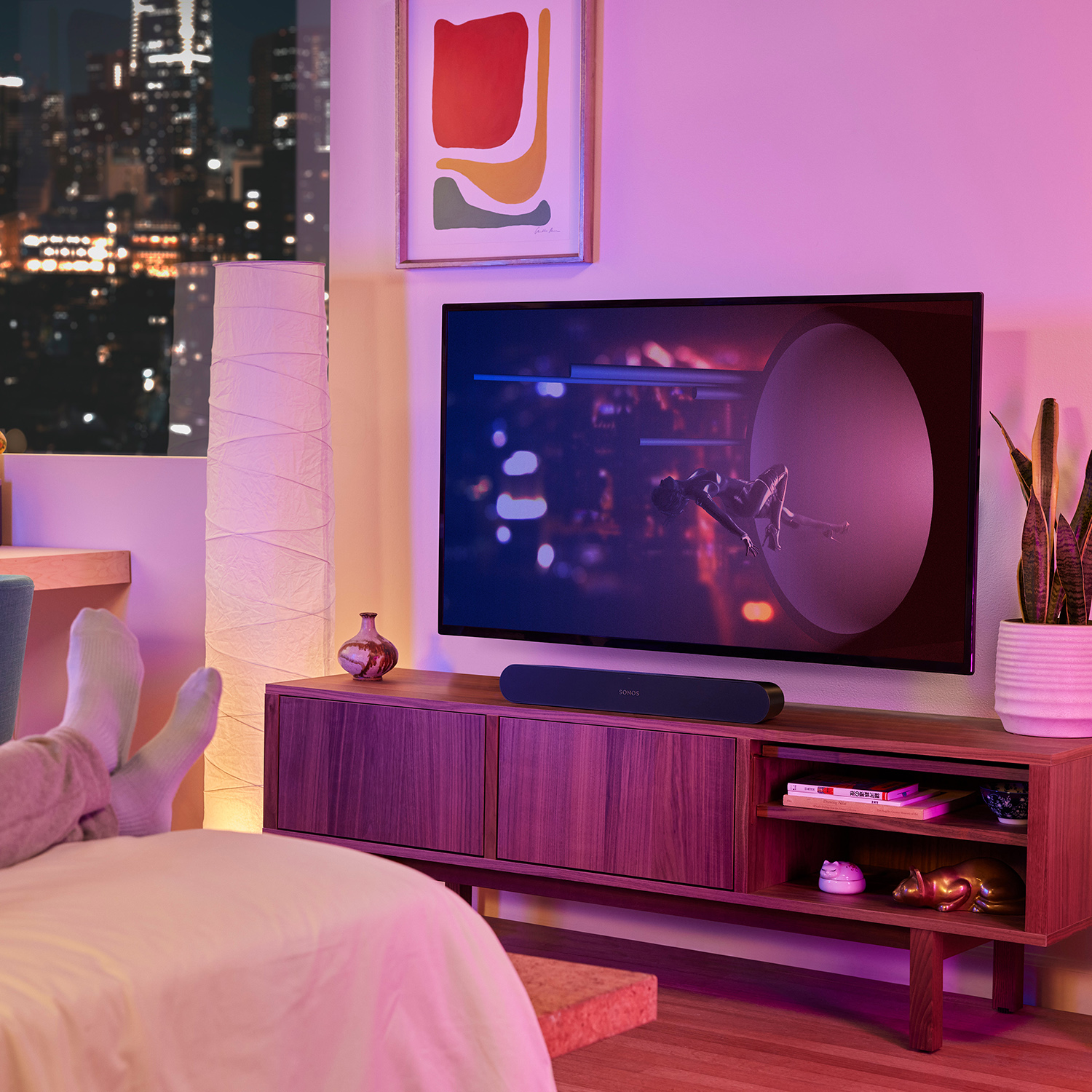 Woonkamerscène. TV, soundbar, meubels en lamp. Voeten rusten op bed. Stad op de achtergrond.