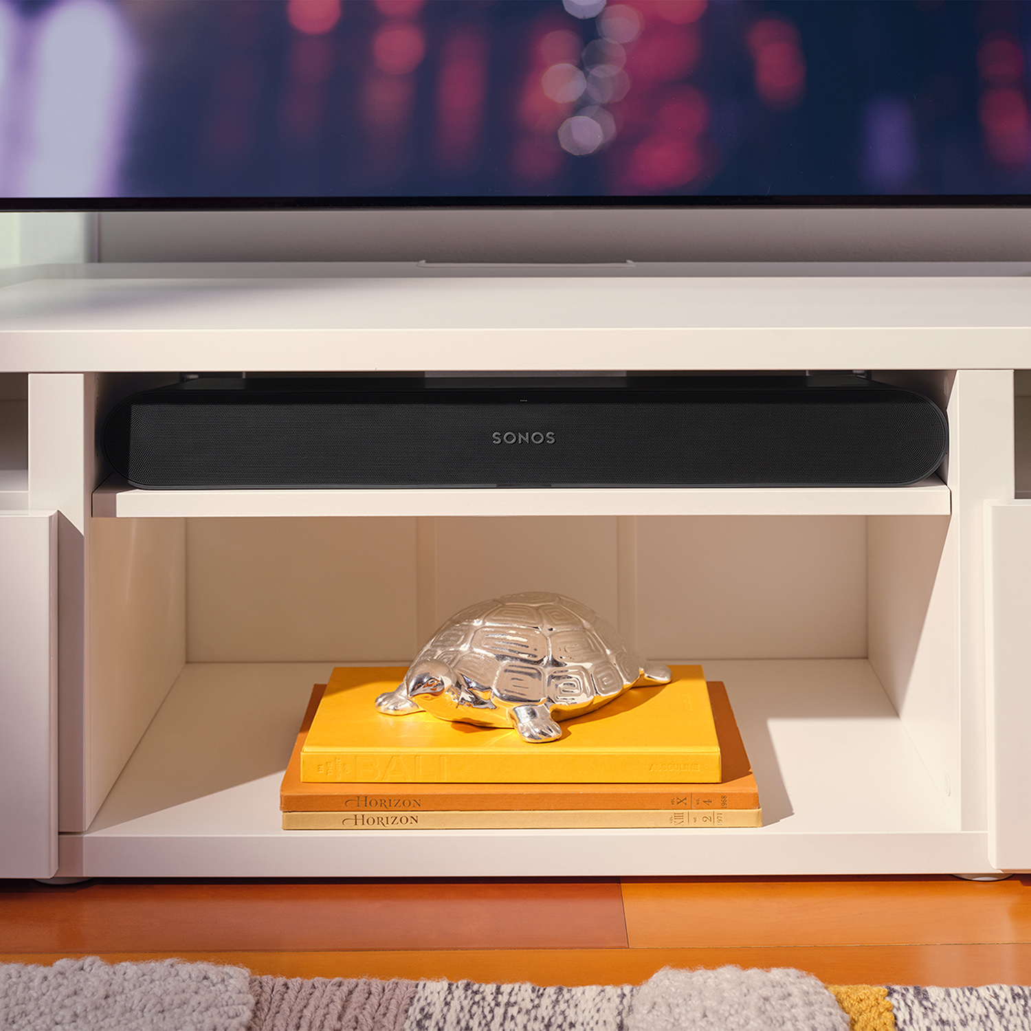 Zwarte soundbar op witte plank. Zilveren schildpad, boeken. TV op de achtergrond.