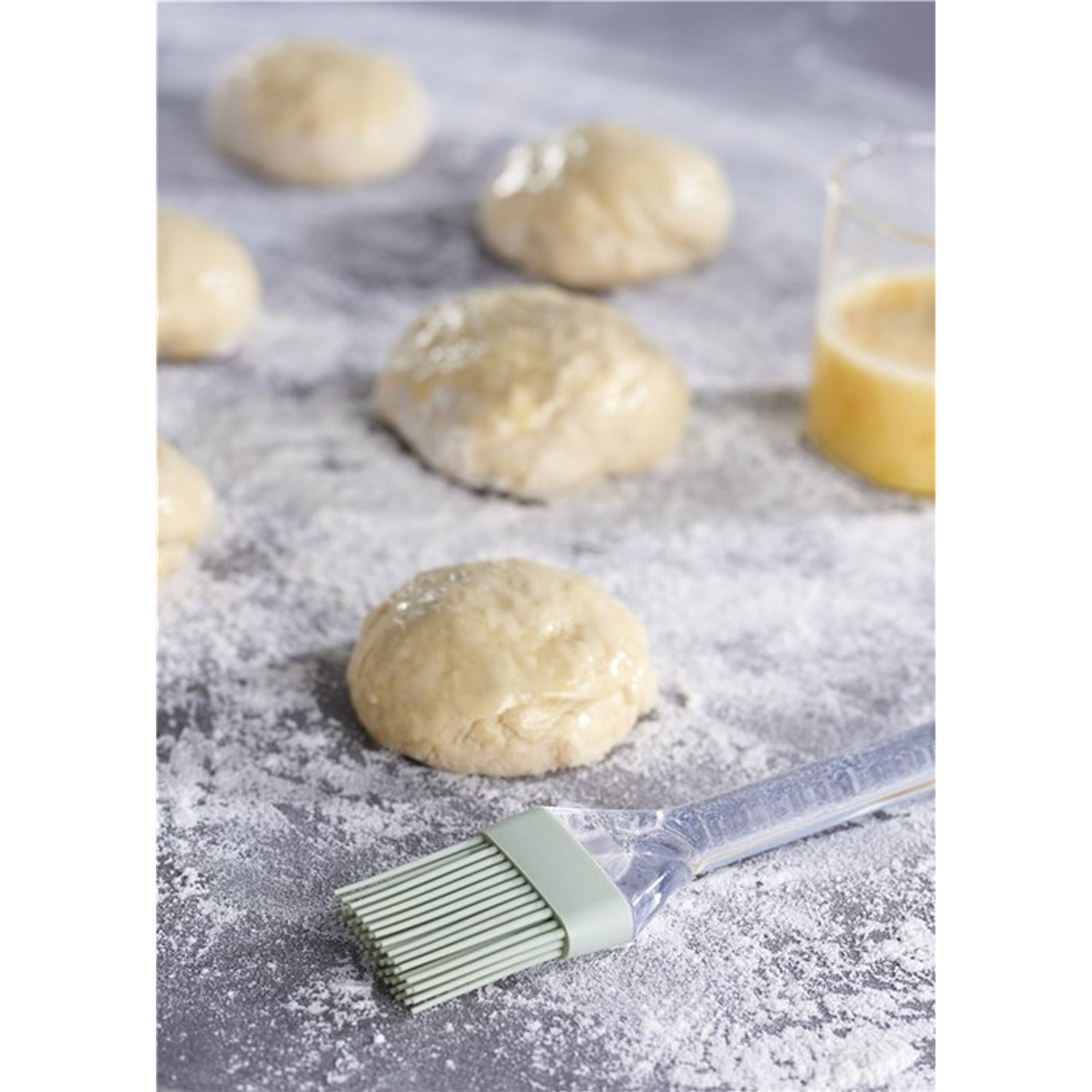 Bolas de masa cruda sobre una superficie enharinada, un pincel con mango verde y un vaso con líquido.