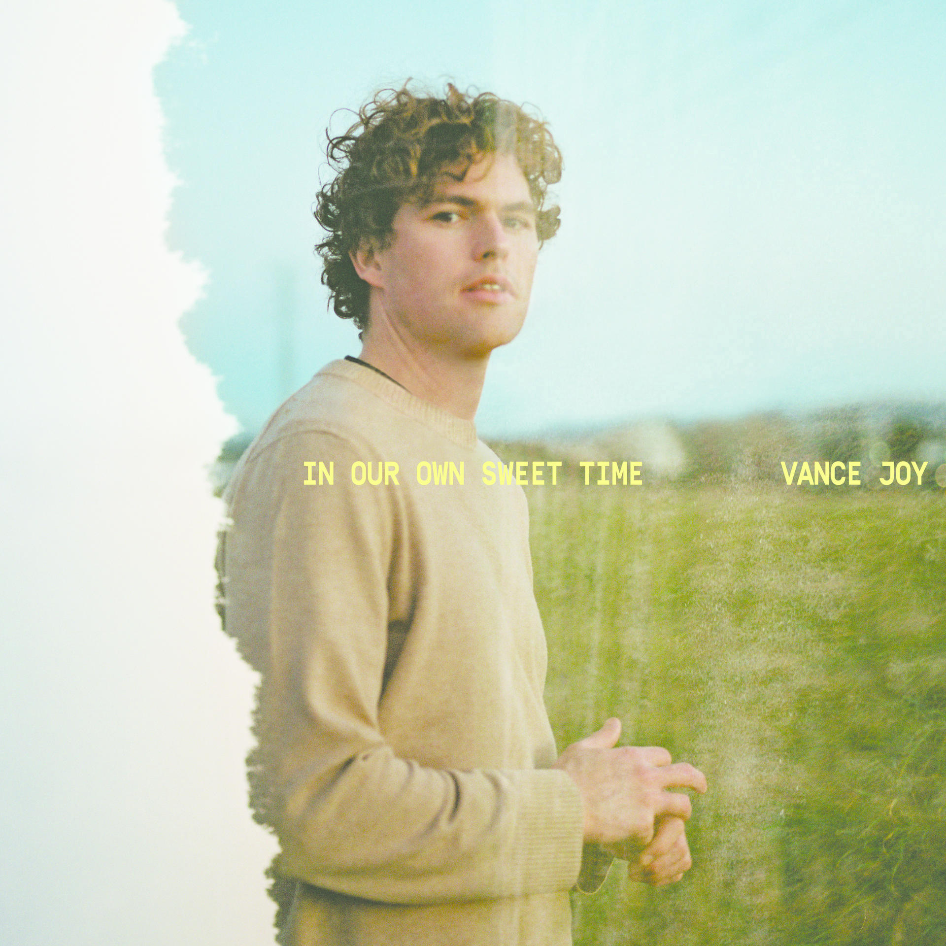 Albumcover mit Vance Joy, beiger Pullover, stehend auf einem Feld, Text "IN OUR OWN SWEET TIME".