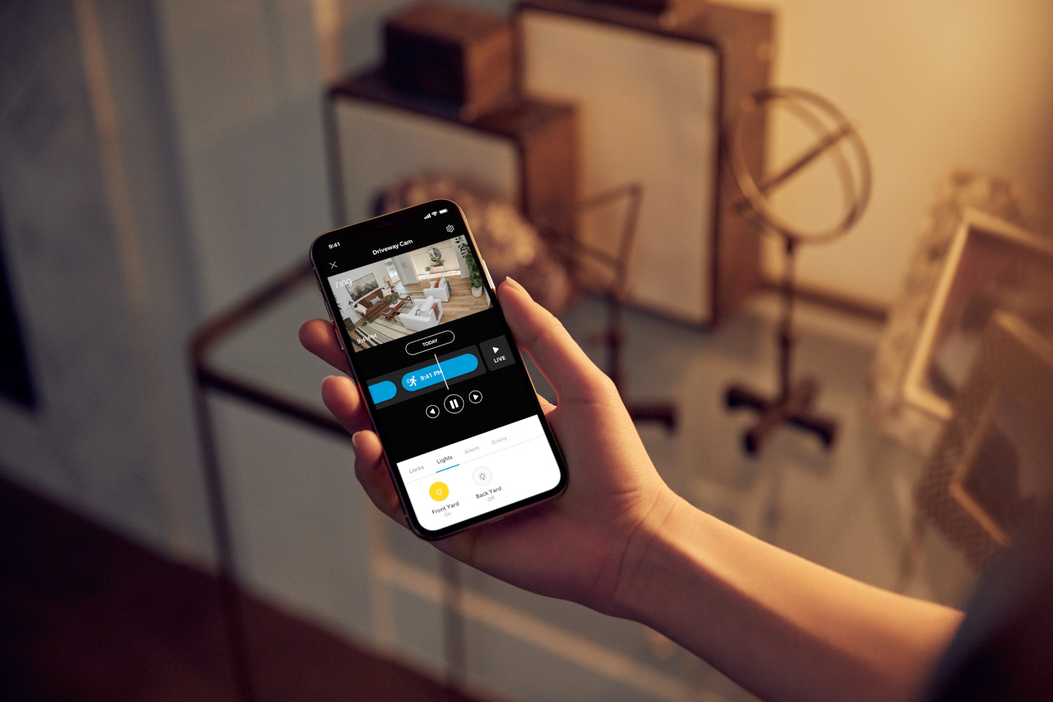 A hand holds a phone displaying a home security camera view. The screen shows a living room. Background items include art and decor.