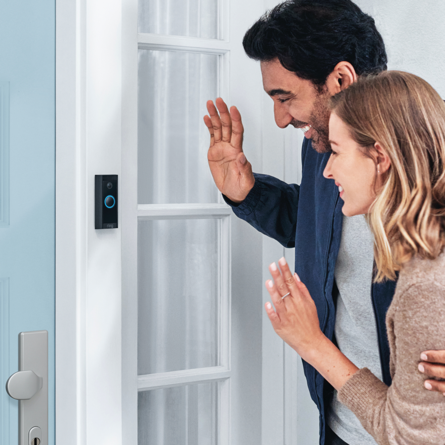 A couple waves at a Ring doorbell camera on a light blue door, standing on the porch.