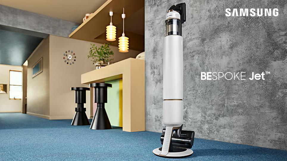 Ein weißer Samsung Bespoke Jet Staubsauger steht auf blauem Teppich. Der Hintergrund zeigt einen Wohnraum.