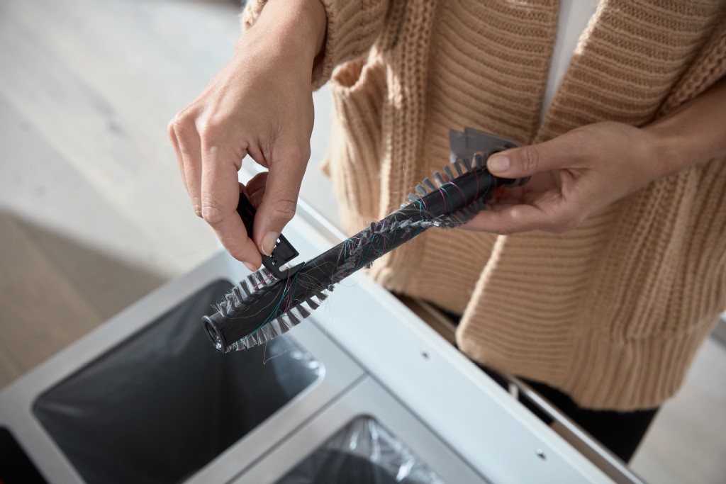 Une personne tenant une brosse d'aspirateur au-dessus d'une poubelle, avec un fond blanc.