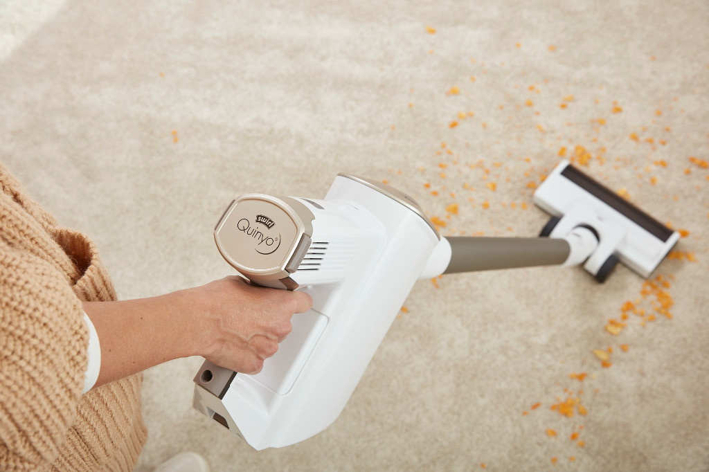 Une personne utilise un aspirateur blanc et beige sur un tapis beige, avec des miettes oranges éparpillées.