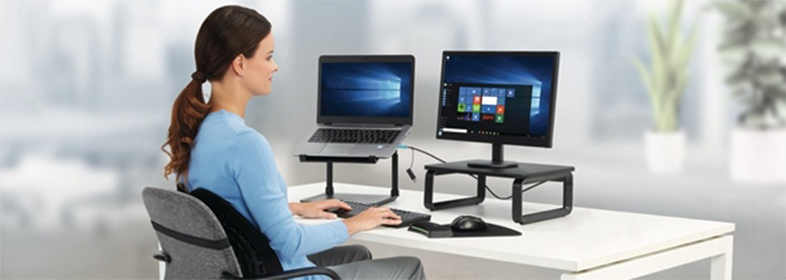 Nő laptopot és monitort használ az asztalnál. Egér, billentyűzet és monitorállvány látható.