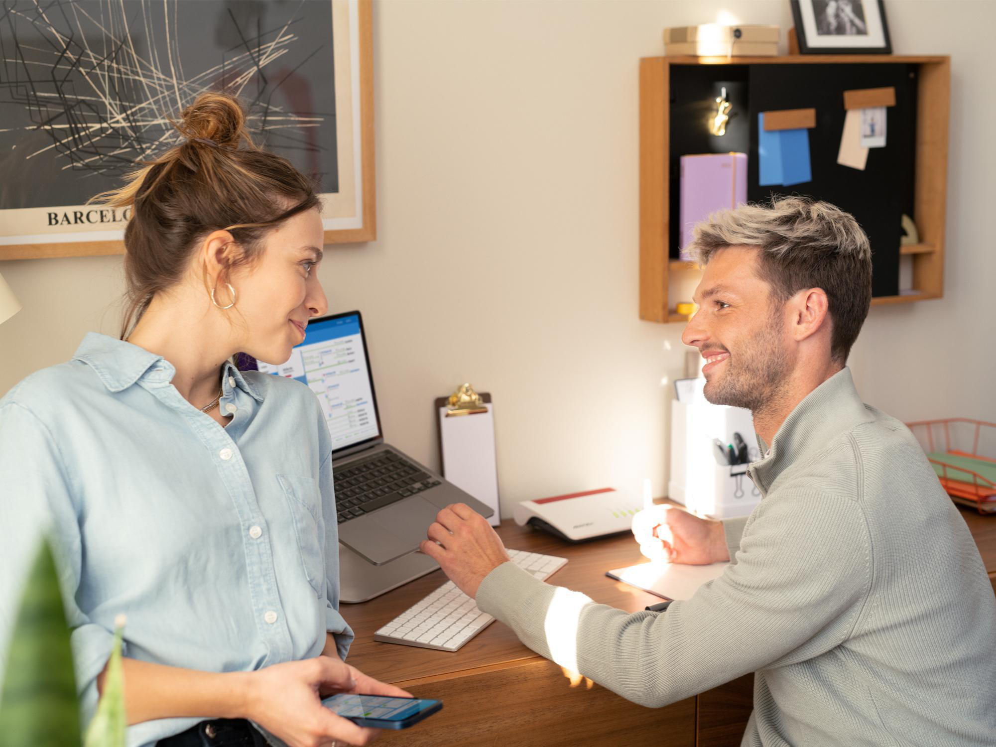 Zwei lächelnde Personen in einem Büro. Frau hält Telefon, Mann beugt sich vor. Laptop und Schreibtisch sichtbar.