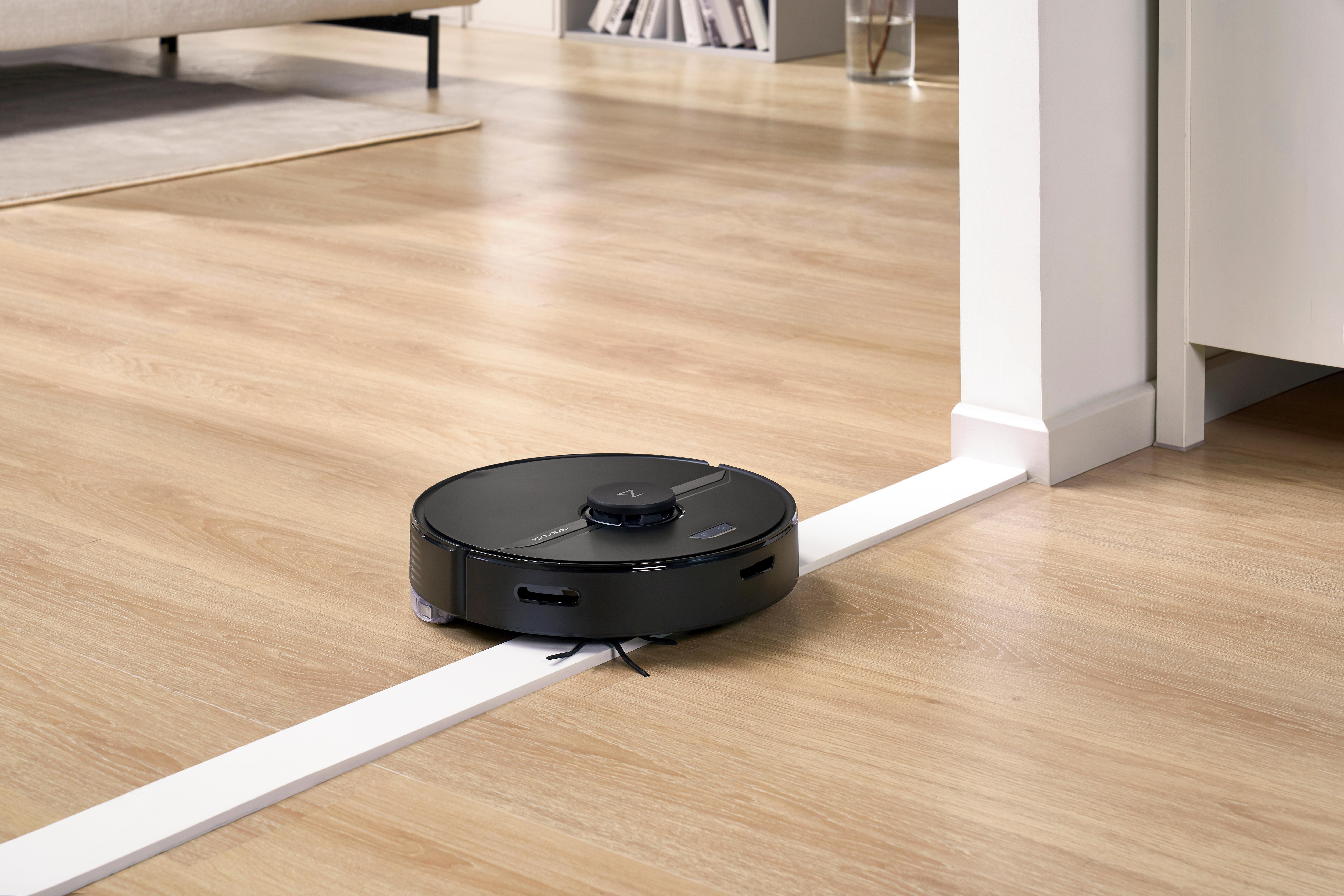 Ein schwarzer Roboter-Staubsauger auf einem Holzboden, der in der Nähe einer weißen Leiste reinigt.