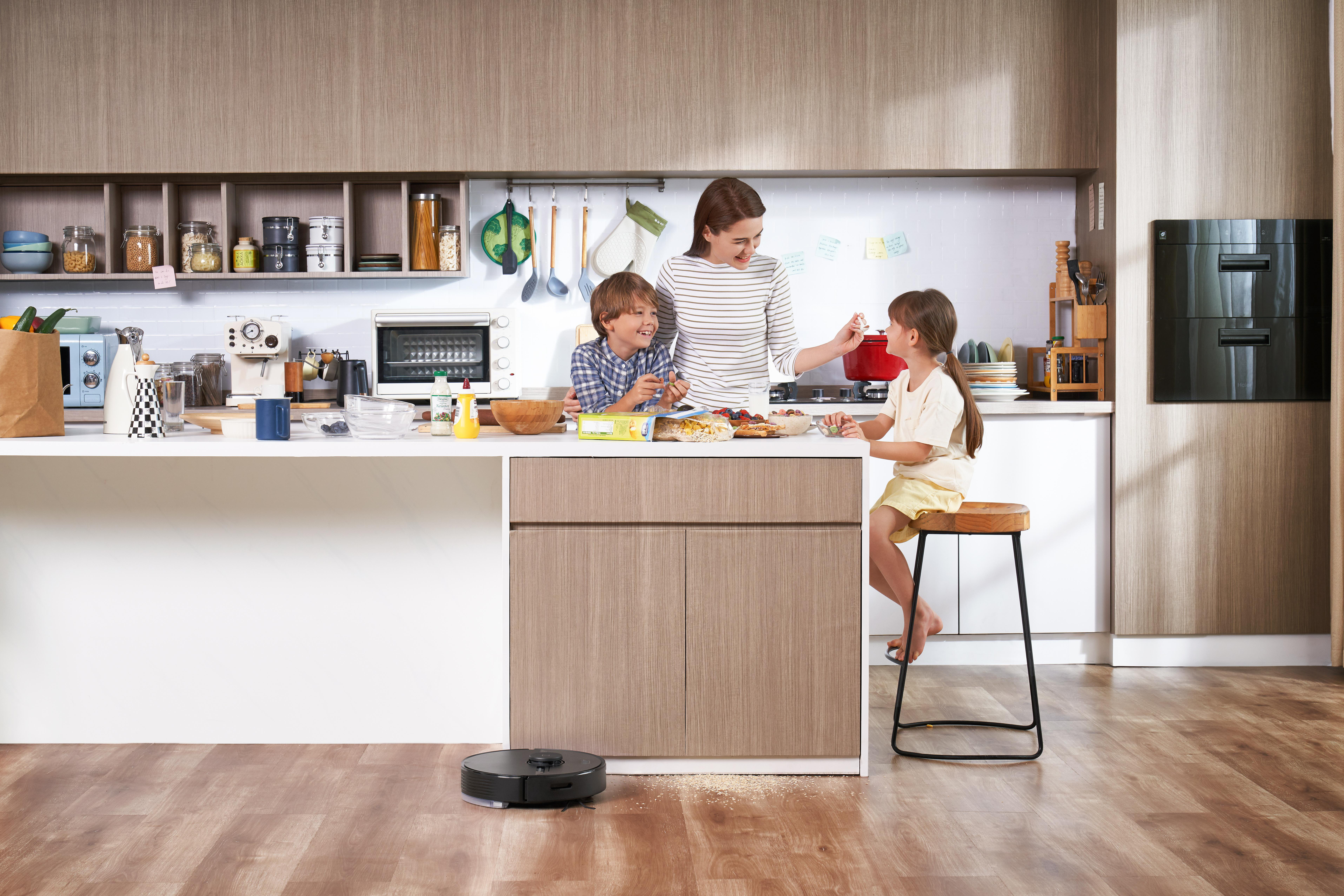 Eine Familie in der Küche bereitet Essen zu, ein Roboterstaubsauger reinigt den Boden.