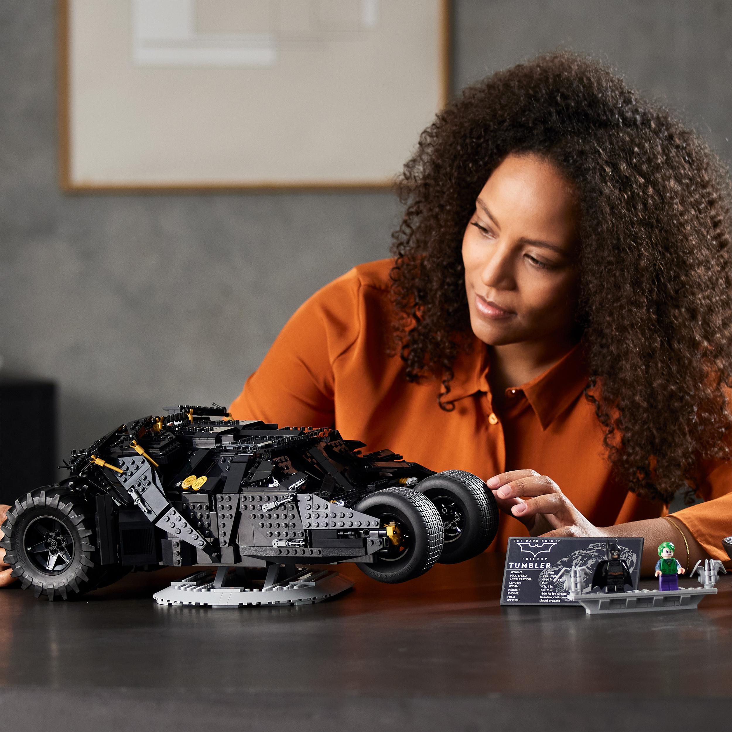Frau mit lockigem Haar betrachtet ein schwarzes Lego Batmobil-Modell auf einem Tisch.