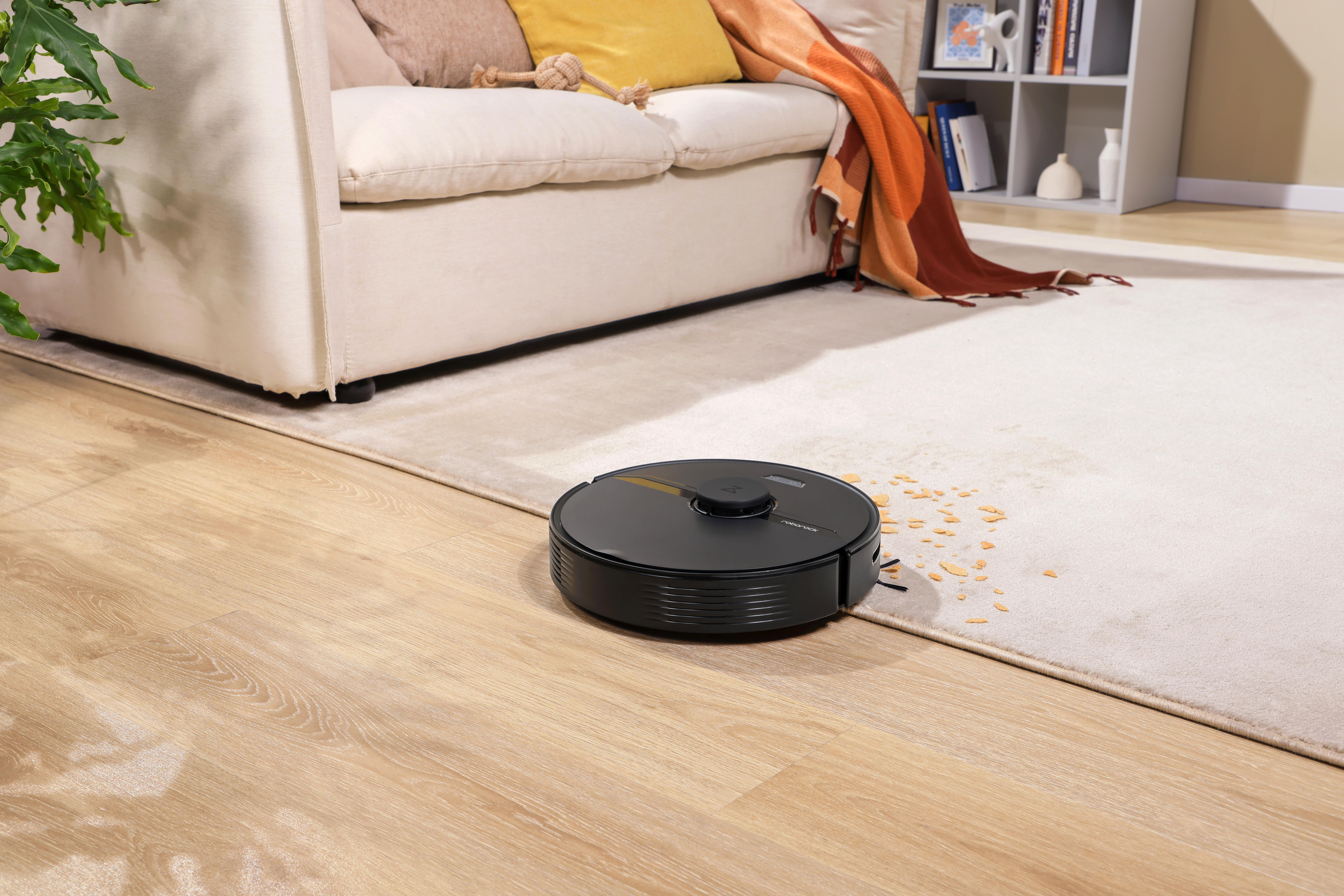 Roboter-Staubsauger auf Teppich mit Schmutz. Sofa und Bücherregal im Hintergrund. Holzboden.
