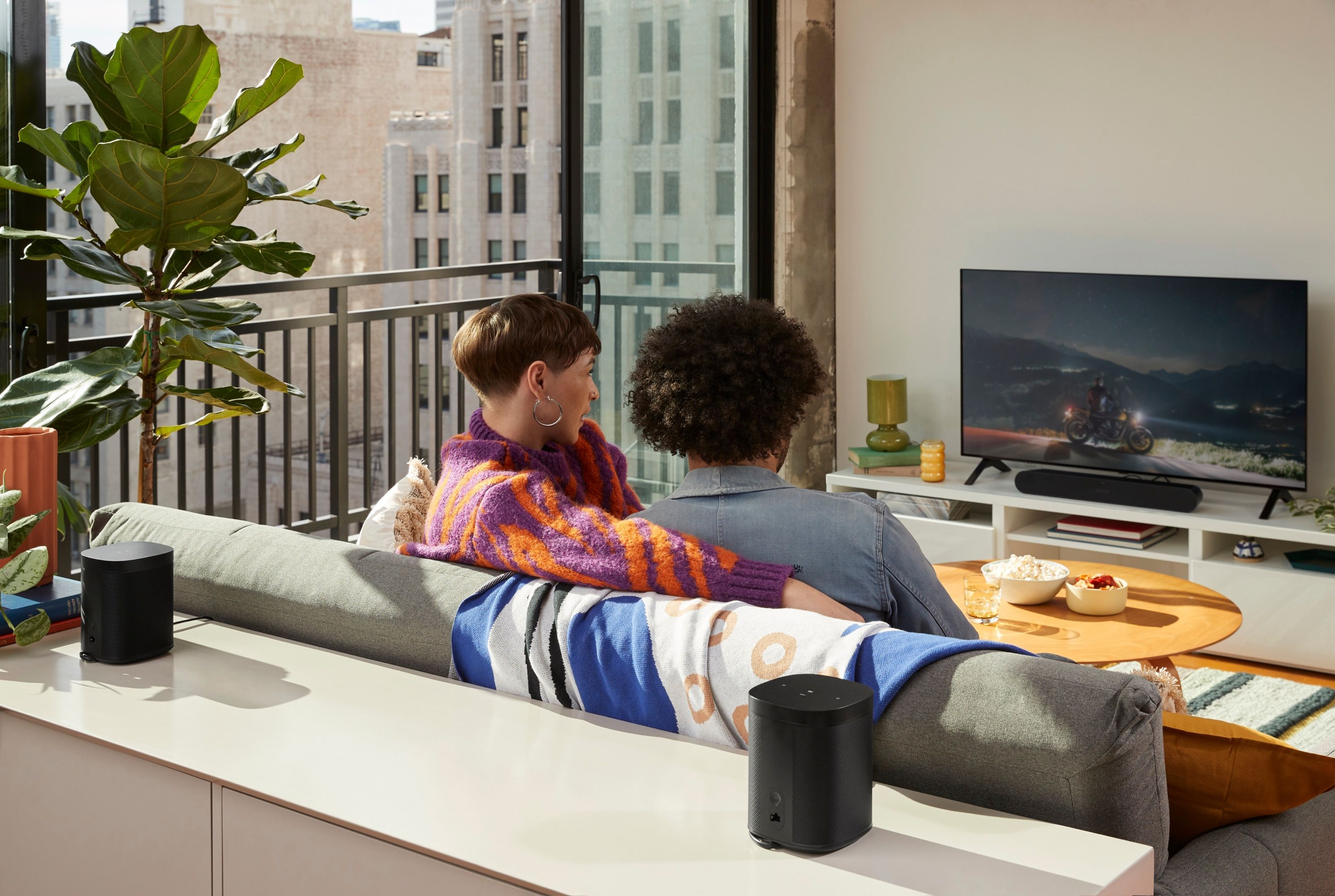 Due persone su un divano che guardano la TV con una soundbar e due altoparlanti neri.